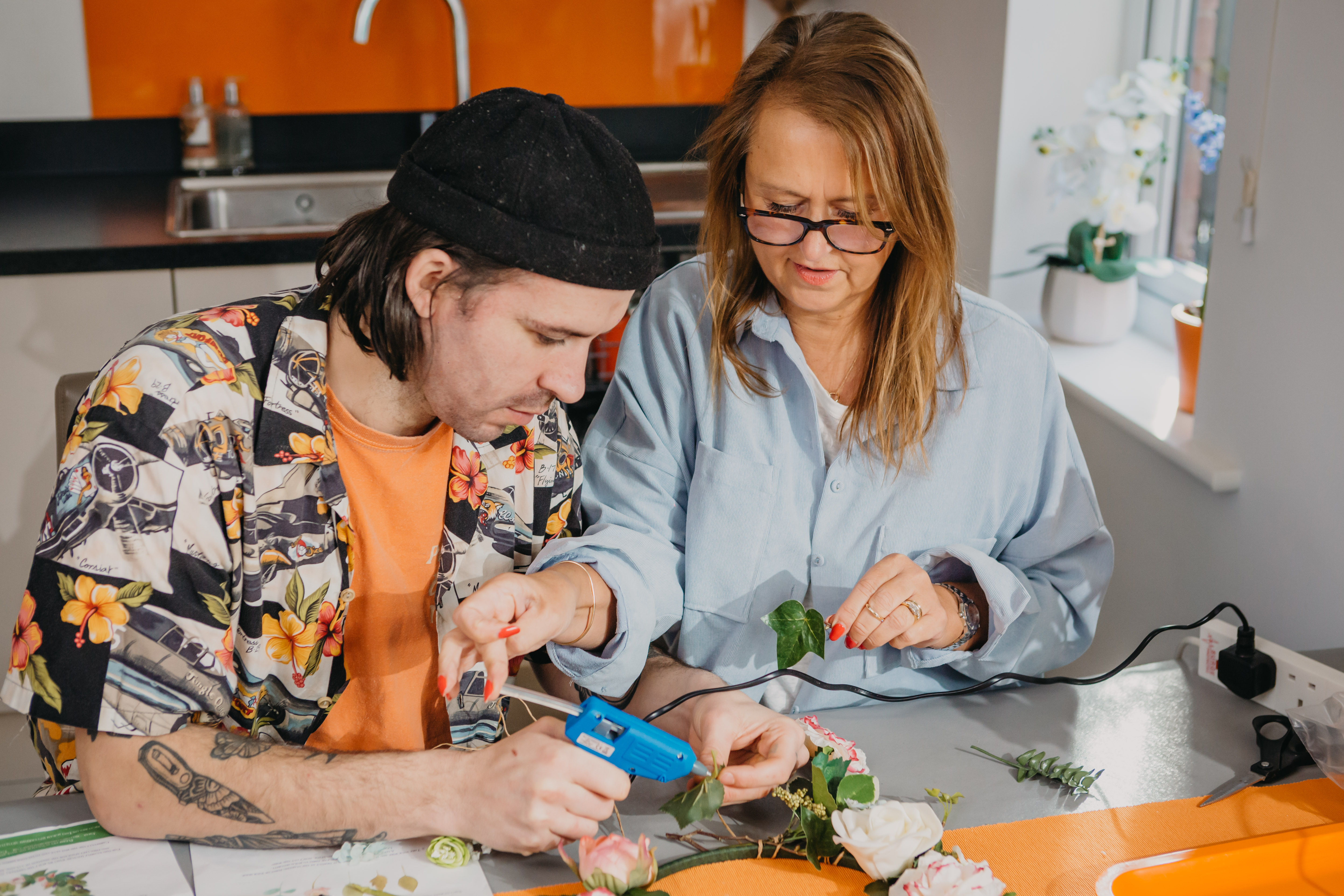 A man in a cap and a lady in glasses make a floral wreath together